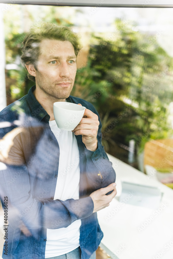 Mid adult man looking through window while drinking coffee standing at home Mid adult man looking through window while drinking coffee standing at home