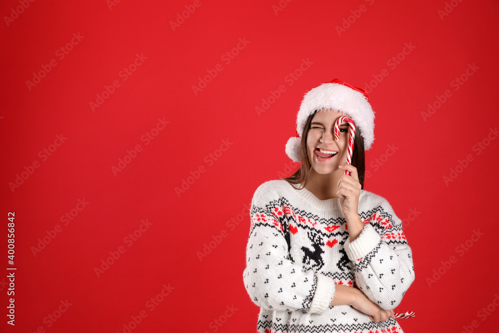 Pretty woman in Santa hat and Christmas sweater holding candy canes on red background, space for text Pretty woman in Santa hat and Christmas sweater holding candy canes on red background, space for text