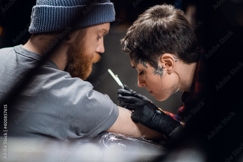A tattoo artist with short hair and piercings is preparing to get a tattoo on a man's arm in a tattoo parlor. A tattoo artist with short hair and piercings is preparing to get a tattoo on a man's arm in a tattoo parlor.