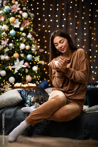 Happy young woman with long hair is holding cup of coffee against bokeh lights, christmas tree. Cozy atmosphere.