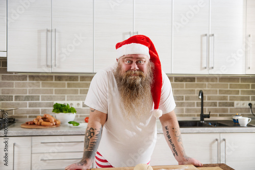 A surprised overweigh man in Santa Claus hat with tattooed arms wearing a white T-Shirt standing in front of the table in the kitchen and looking at camera.  photo