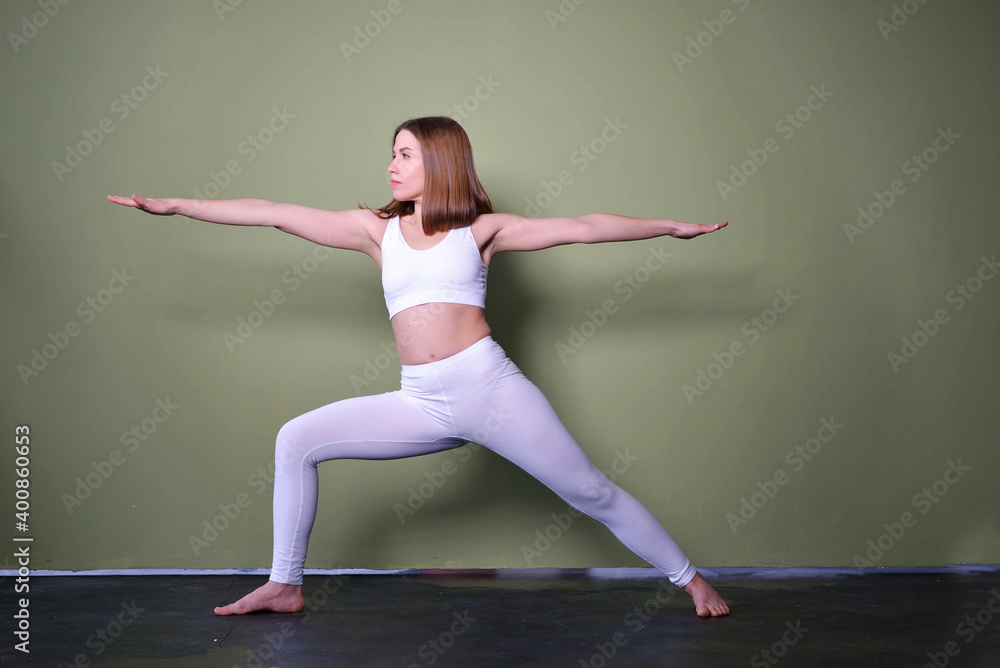 Young attractive woman practicing yoga, standing in Warrior one exercise, Virabhadrasana II pose, working out, wearing sportswear, indoor full length, studio background.