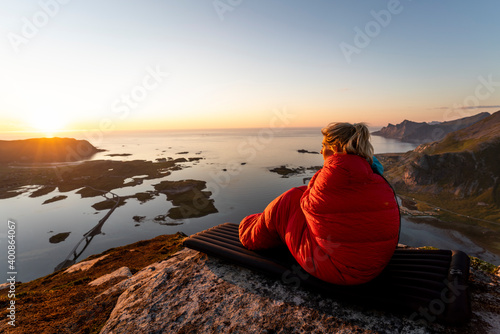 Woman wrapped in sleeping bag looking at view
while sitting on mountain at Volandstinden, Lofoten, Norway photo