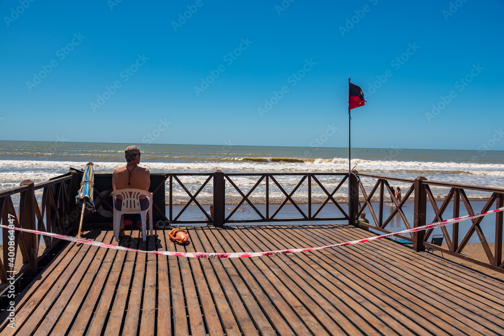 Mar del Tuyú, Buenos Aires, Argentina; December 06, 2020: Lifeguard sitting on a deck next to the tidal flag. A do not cross link because social distancing during COVID-19