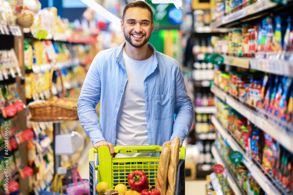 Young cheerful bearded man shopping in supermarket with trolley cart