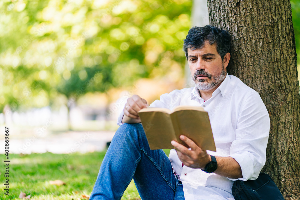 Mature man reading book while leaning on tree trunk in public park Mature man reading book while leaning on tree trunk in public park