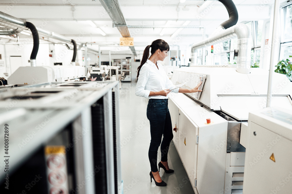 Businesswoman examining machinery while holding digital tablet in industry Businesswoman examining machinery while holding digital tablet in industry