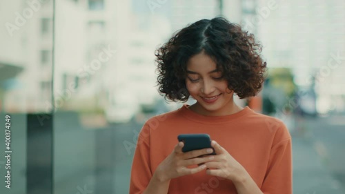 Attractive Young Indian woman in casual clothes walking in a city street outdoors looking at mobile phone.  Smiling Girl using a smartphone browsing internet, searching, reading news in social media.