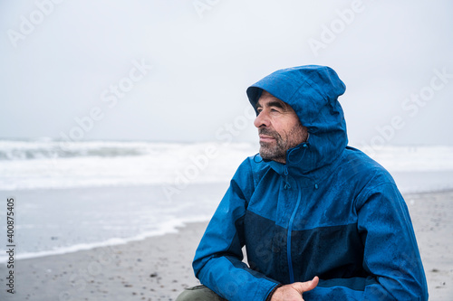 Contemplated mature man in blue raincoat crouching at beach against sky photo