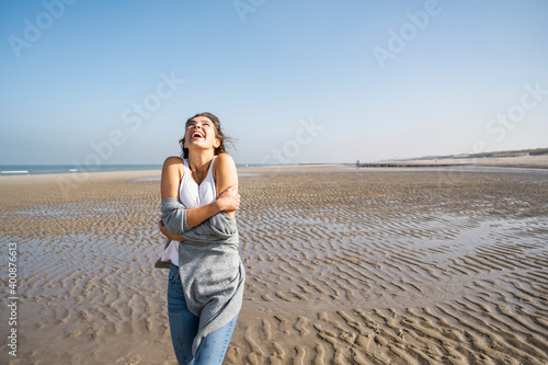 Happy young woman hugging self while standing at beach against clear sky photo