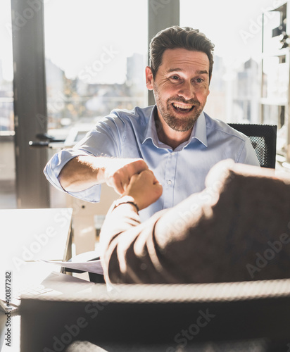 Happy businessman giving fist bump to colleague while sitting at office
