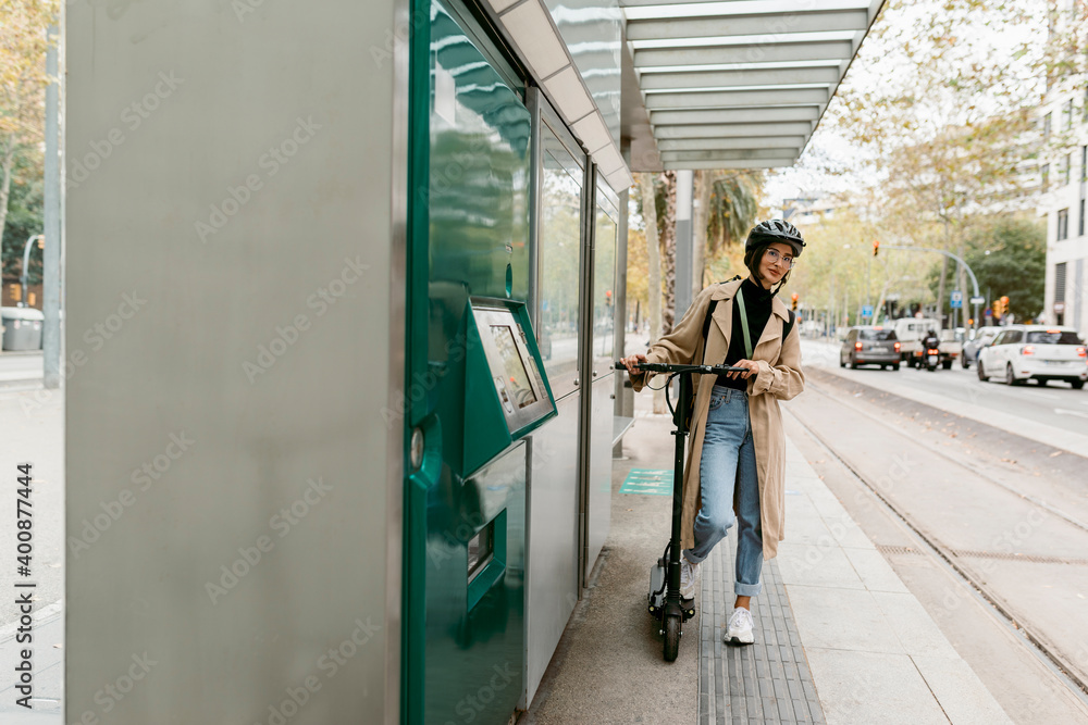 Woman wearing cycling helmet standing with electric push scooter while waiting at tram station in city Woman wearing cycling helmet standing with electric push scooter while waiting at tram station in city