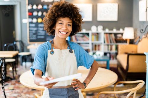Happy female owner giving menu while standing with hand on hip at coffee shop
