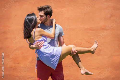 Young man kissing while carrying woman against brown wall at back yard during sunny day © Javier Sanchez Mingorance/Westend61