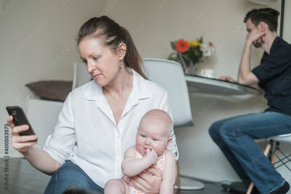 Mother holding baby (2-5 months) while using smart phone and father working in background Mother holding baby (2-5 months) while using smart phone and father working in background