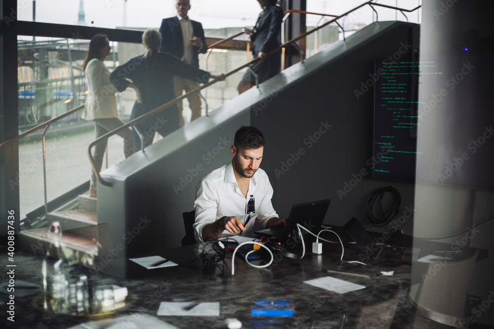 Business professional working on laptop while coworkers discussing on staircase in creative office Business professional working on laptop while coworkers discussing on staircase in creative office