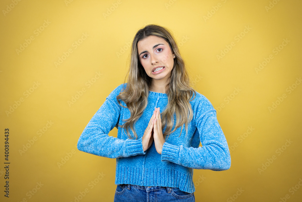 Pretty blonde woman with long hair standing over yellow background begging and praying with hands together with hope expression on face very emotional and worried Pretty blonde woman with long hair standing over yellow background begging and praying with hands together with hope expression on face very emotional and worried