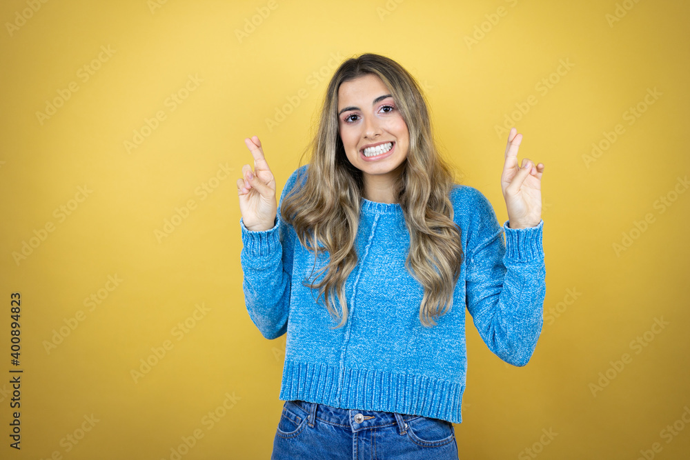 Pretty blonde woman with long hair standing over yellow background gesturing finger crossed smiling with hope and looking side Pretty blonde woman with long hair standing over yellow background gesturing finger crossed smiling with hope and looking side