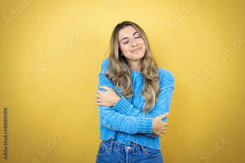 Pretty blonde woman with long hair standing over yellow background hugging oneself happy and positive, smiling confident