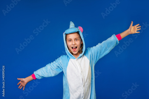 Charming girl playfully poses in the costume of the unicorn. A Studio shot of an emotional woman in kigurumi having fun on a blue background. Cheerfully spreading his hands to the sides.