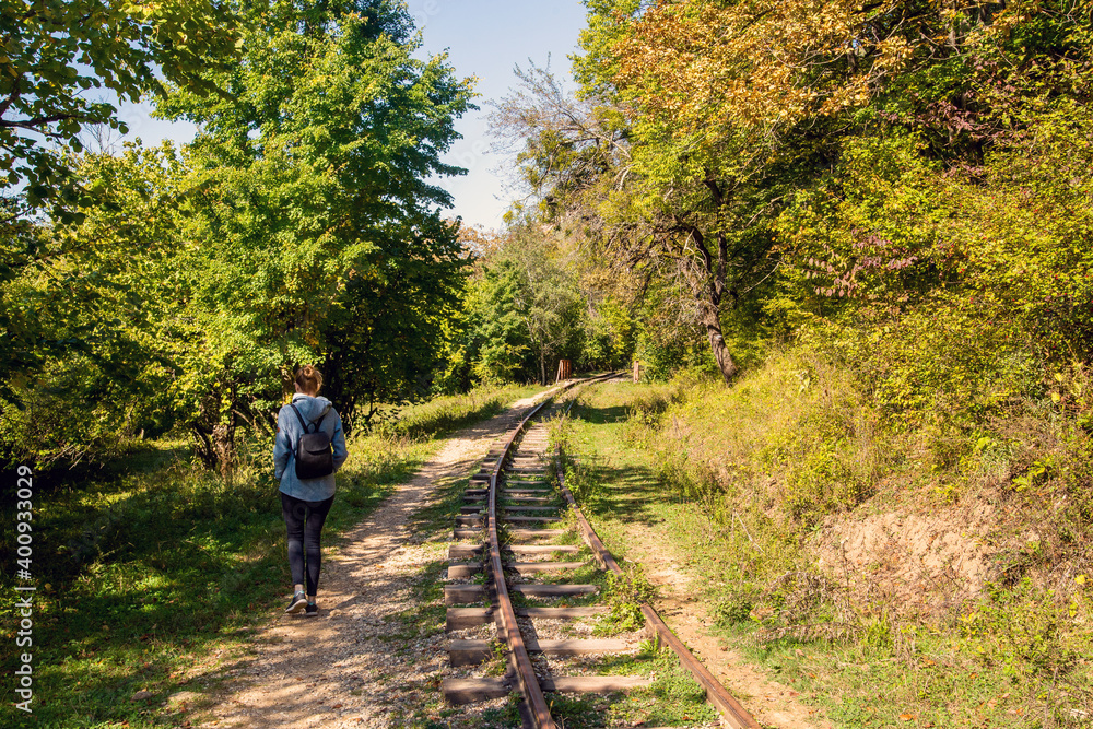 Naklejka premium Rear view of a young hiking tourist girl enjoying amazing beauty of nature. Excursion on old narrow railroad in forest