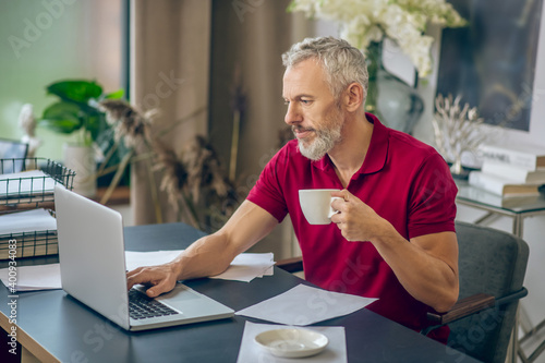 Grey-haired man sitting at the laptop and having coffee