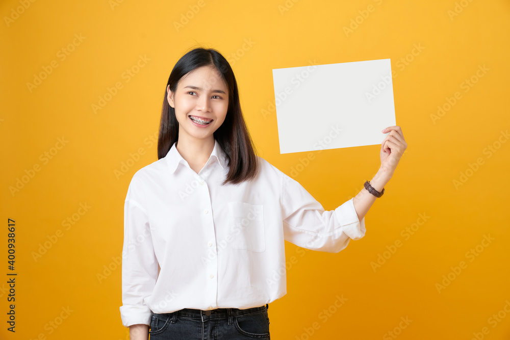 Young Asian woman holding blank paper with smiling face and looking on the yellow background. for advertising signs. Young Asian woman holding blank paper with smiling face and looking on the yellow background. for advertising signs.