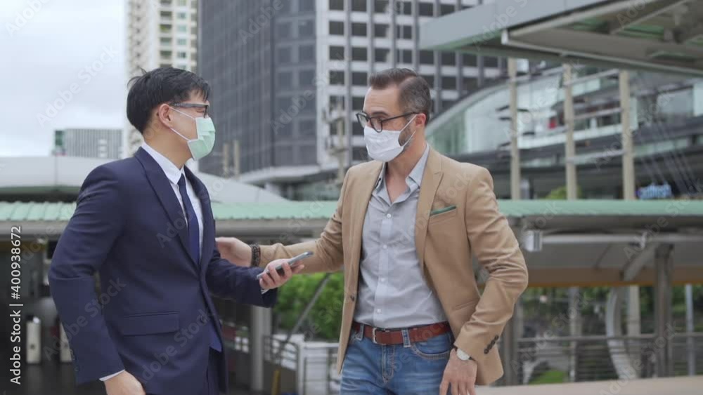 Business people Caucasian with Asian man office worker colleague partnership wearing protective face mask for protect covid-19 virus discussing business project plan with using smartphone in the city Business people Caucasian with Asian man office worker colleague partnership wearing protective face mask for protect covid-19 virus discussing business project plan with using smartphone in the city