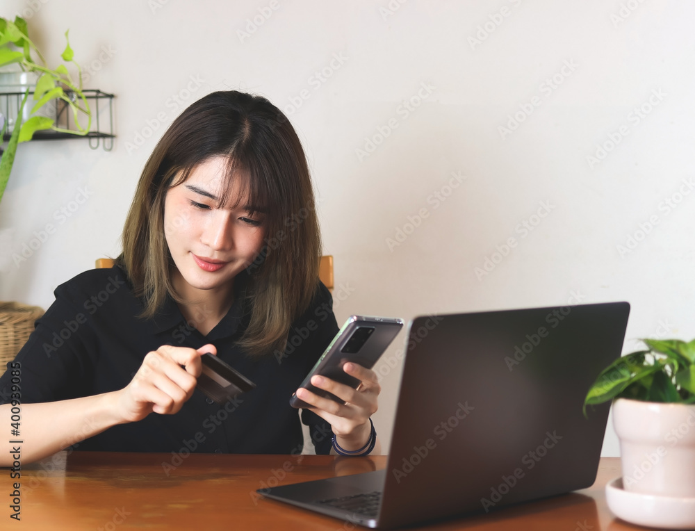 Asian woman wearing black shirt, sitting at wooden table with computer laptop, holding credit card and mobile phone. white wall with house plant background. online shopping concept. Asian woman wearing black shirt, sitting at wooden table with computer laptop, holding credit card and mobile phone. white wall with house plant background. online shopping concept.