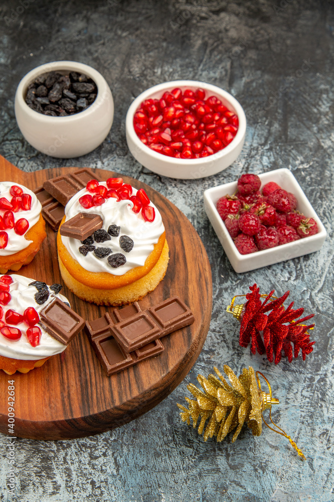 top view berry cakes and chocolates on chopping board xmas toys bowls with pomegranate raisins and raspberries on grey-white background top view berry cakes and chocolates on chopping board xmas toys bowls with pomegranate raisins and raspberries on grey-white background