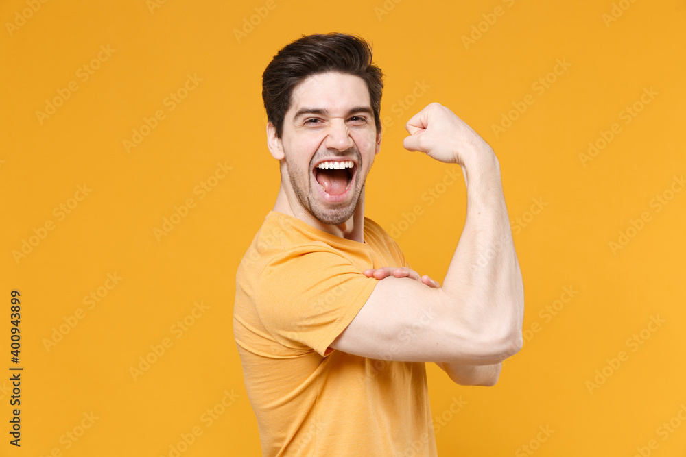 Obraz premium Side view of young strong muscular smiling unshaved caucasian handsome man 20s years old in casual basic blank print design t-shirt showing biceps muscles isolated on yellow background studio portrait