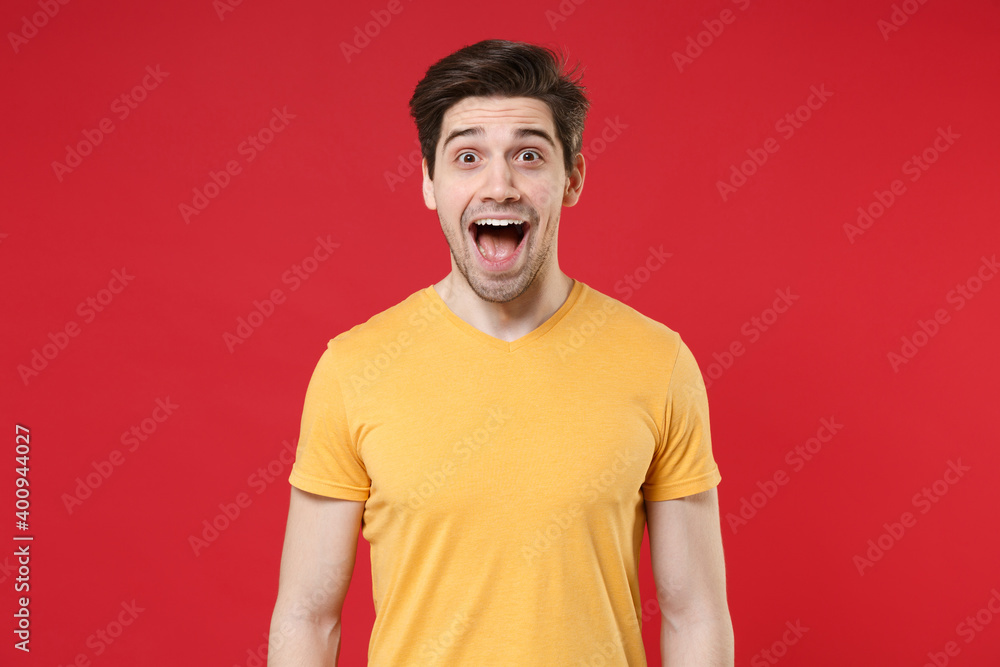 Young surprised shocked excited unshaved caucasian satisfied wonder man 20s wearing casual basic blank print design yellow t-shirt with looking camera isolated on red color background studio portrait. Young surprised shocked excited unshaved caucasian satisfied wonder man 20s wearing casual basic blank print design yellow t-shirt with looking camera isolated on red color background studio portrait.