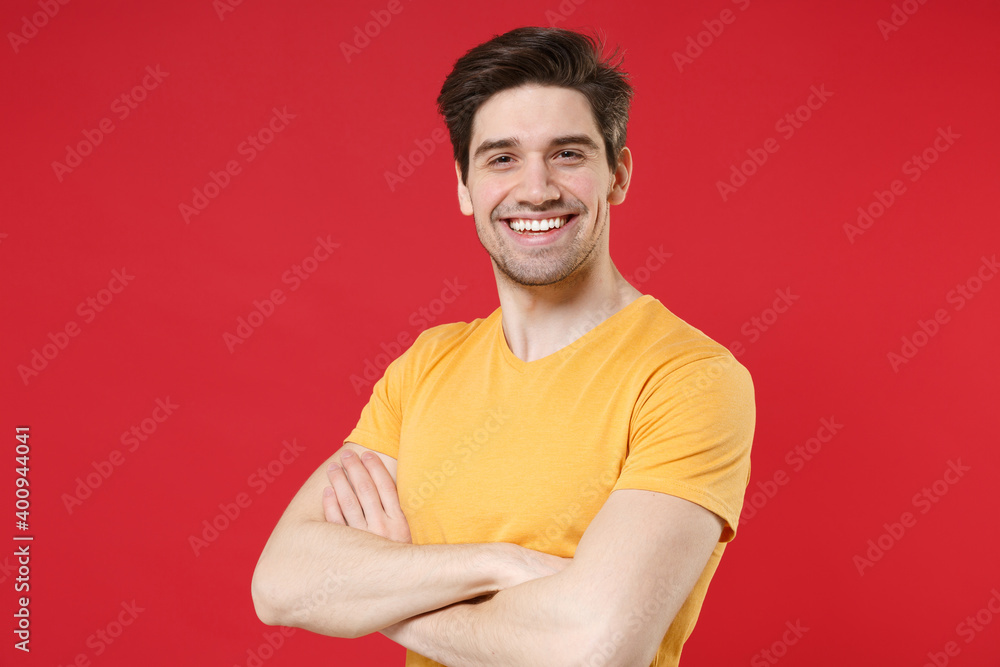 Obraz premium Young smiling unshaved cheerful friendly caucasian handsome man 20s wearing casual basic yellow t-shirt looking camera holding hands crossed folded isolated on red color background studio portrait.