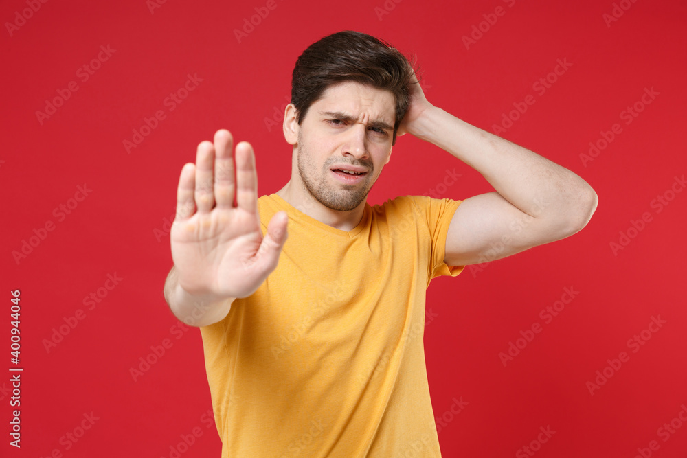 Young refusing sick exhausted depressed unshaved tired caucasian man 20s in casual yellow t-shirt showing stop palm gesture put hand forehead head isolated on red color background studio portrait. Young refusing sick exhausted depressed unshaved tired caucasian man 20s in casual yellow t-shirt showing stop palm gesture put hand forehead head isolated on red color background studio portrait.