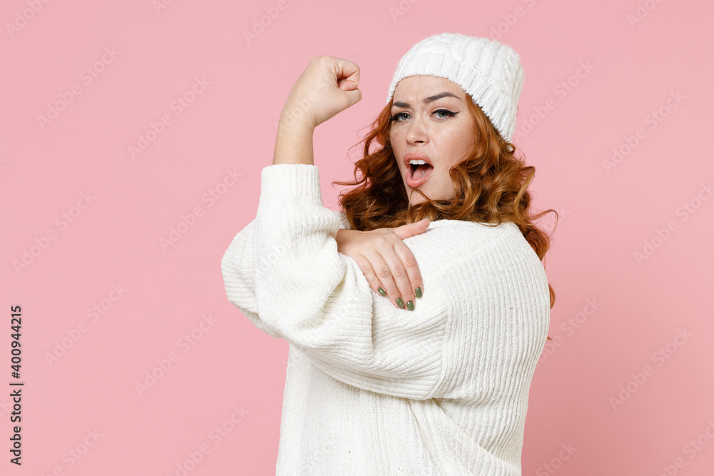 Side view of strong young redhead plus size body positive woman 20s in warm white sweater hat standing showing biceps muscles looking camera isolated on pastel pink color background studio portrait. Side view of strong young redhead plus size body positive woman 20s in warm white sweater hat standing showing biceps muscles looking camera isolated on pastel pink color background studio portrait.