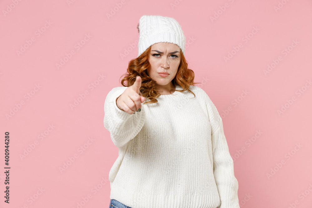 Displeased strict young redhead plus size body positive woman 20s in casual warm white sweater hat standing pointing index finger on camera isolated on pastel pink color background studio portrait. Displeased strict young redhead plus size body positive woman 20s in casual warm white sweater hat standing pointing index finger on camera isolated on pastel pink color background studio portrait.