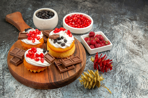 bottom view berry cakes and chocolates on chopping board colorful pinecones bowls with pomegranate raisins and raspberries on grey-white background