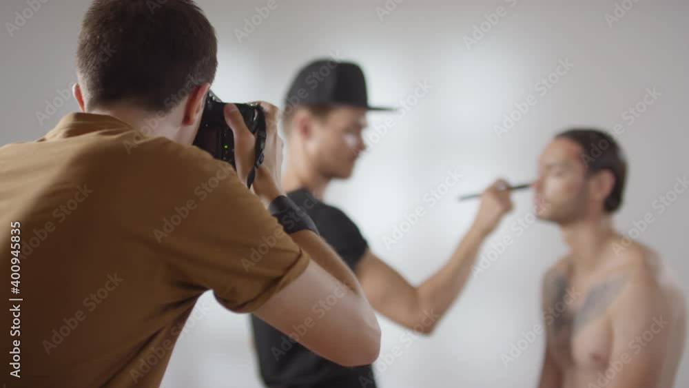 Medium shot of photographer doing photo shoot of professional male beautician while he is applying make-up on client Medium shot of photographer doing photo shoot of professional male beautician while he is applying make-up on client