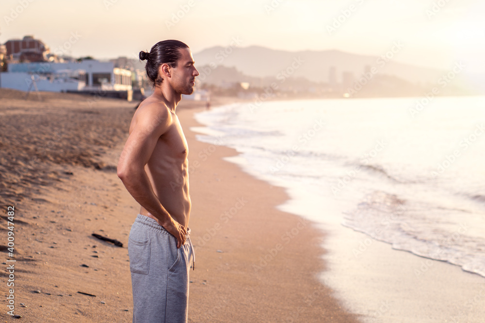 Fitness man standing on the beach without shirt. Athletic fit male shirtless. Showing abs. Personal trainer posing.