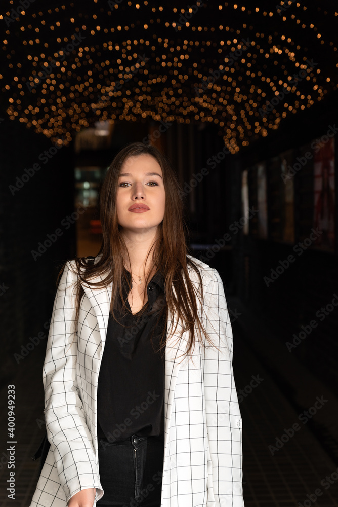 Fototapeta premium Portrait of brunette girl on evening street and garlands background. Vertical frame