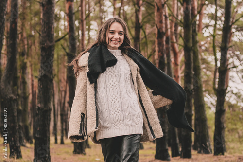 a woman of model appearance with long dark hair holds a scarf in her hands and smiles while in a pine forest
