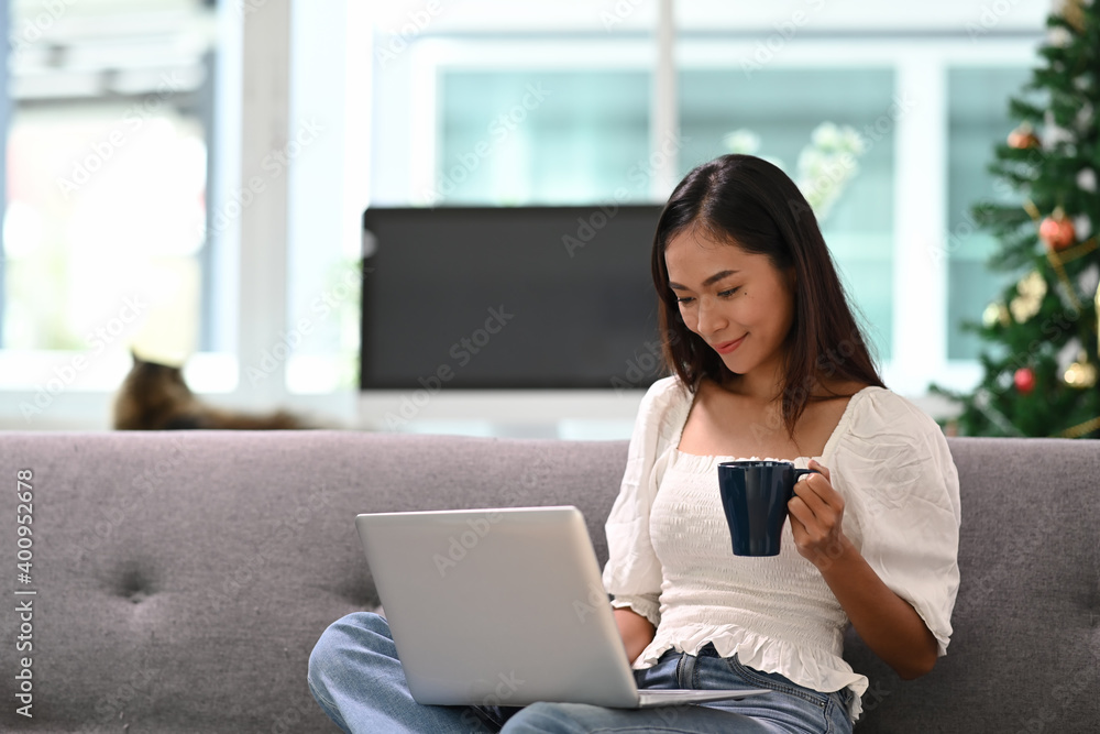 Cheerful young woman using laptop watching funny video and drinking coffee while sitting on couch in living room. Cheerful young woman using laptop watching funny video and drinking coffee while sitting on couch in living room.