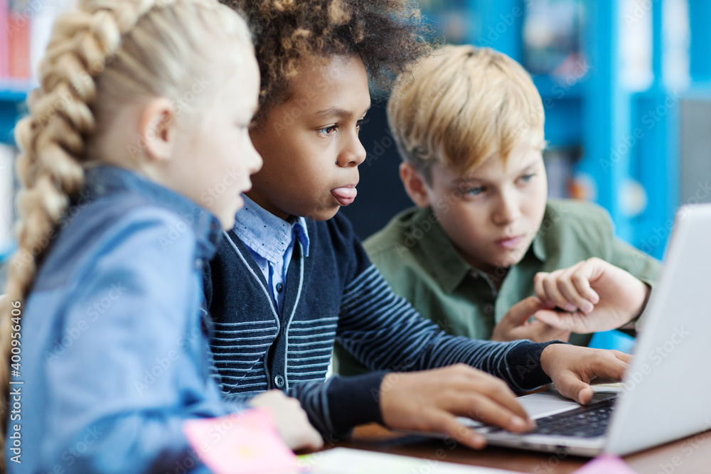 Side view of three curious diverse students with laptop sitting at desk in elementary school classroom. Mixed  race schoolboy and his friends searching for information on Internet