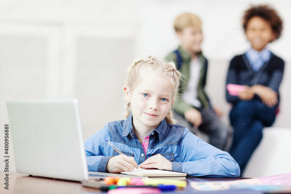 Diligent little girl looking at camera and smiling slightly while writing in notepad sitting at desk in school classroom with open laptop in front of her, two defocused classmates laughing Diligent little girl looking at camera and smiling slightly while writing in notepad sitting at desk in school classroom with open laptop in front of her, two defocused classmates laughing