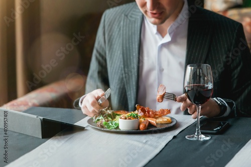 A young and attractive man uses his tablet while eating Traditional English breakfast. Sausage  beans  eggs  bread and salad in an indoor restaurant.