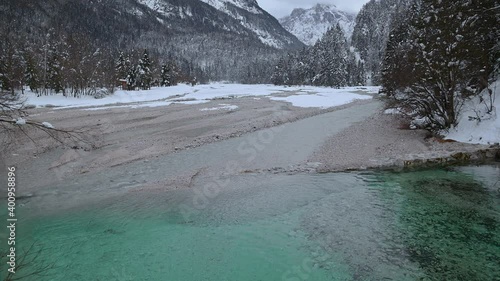 Small narrow green river Pisnica near Kranjska Gora, Slovenia, in winter season. Alps mountains covered with snow in wintertime. Idyll and pristine nature. Static shot, real time, wide angle photo