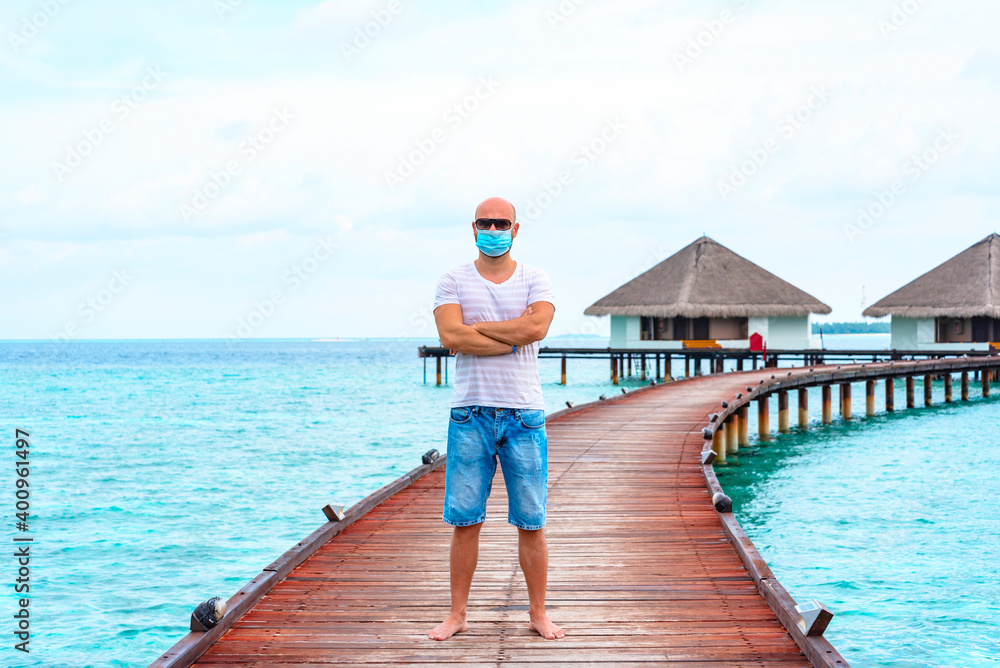 a young man wearing a protective face mask stands on a pier near water villas in the Maldives, a concept of  travel during the covid pandemic