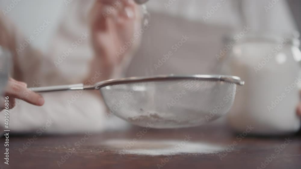 Midsection slow-motion close-up of unrecognizable female hands sifting white flour at kitchen table using sieve and tablespoon Midsection slow-motion close-up of unrecognizable female hands sifting white flour at kitchen table using sieve and tablespoon