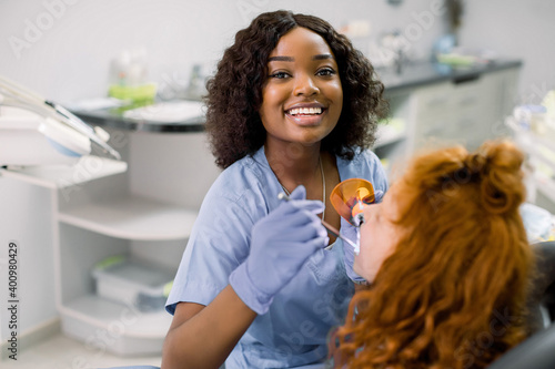 Smiling black woman dentist wearing blue uniform, providing tooth restoration and filling with curing polymerization UV lamp for her little patient, cute girl with red curly hair