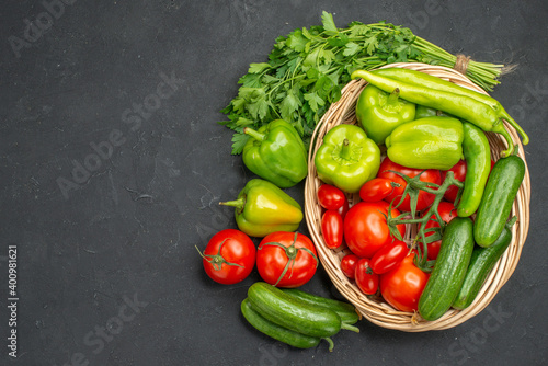 Horizontal view of fresh vegetables red tomatoes with stems green peppers and cucumbers inside and outside of basket on dark background